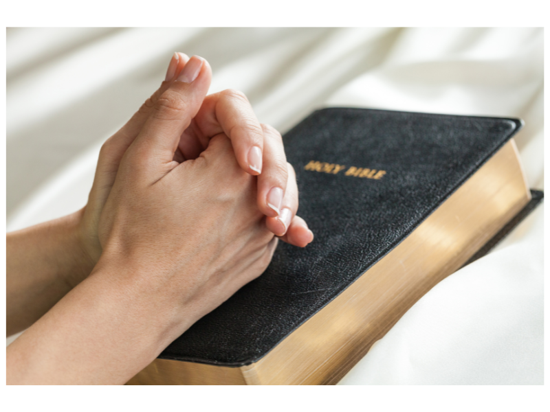 Shows a mother's hands folded in prayer over a Bible.