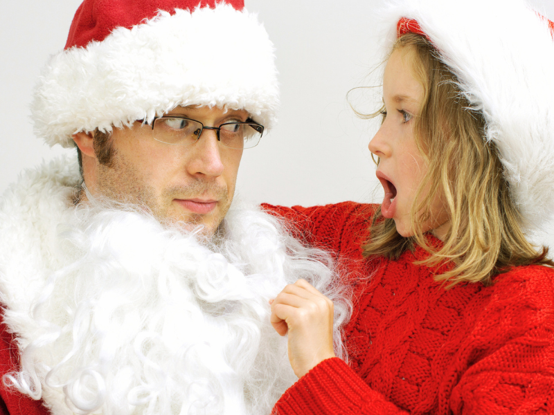 Surprised young girl pulling fake beard away from face of man dressed in a Santa suit.